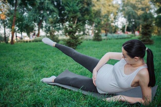 Giovane donna incinta seria che allunga le gambe sul compagno di yoga in parco. Tiene una mano sulla pancia. Le gambe giuste sono in aria.