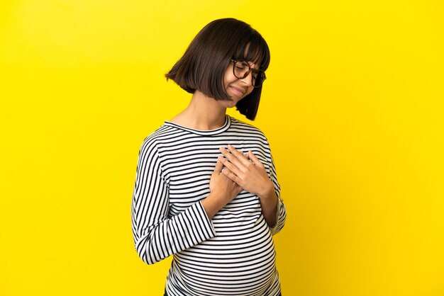 Giovane donna incinta sopra la parete gialla isolata che ha un dolore nel cuore
