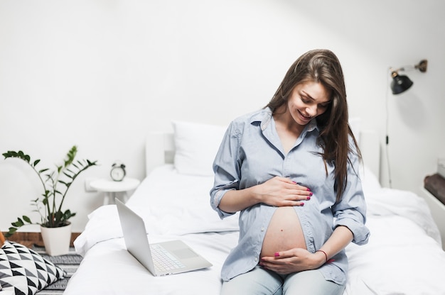 Giovane donna incinta sorridente che si siede vicino al computer portatile sul letto che tiene la sua pancia
