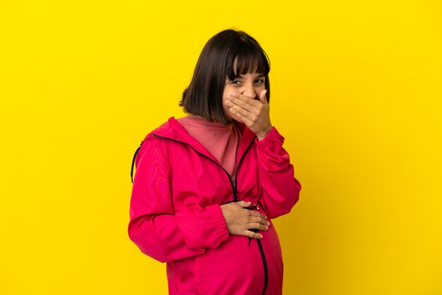 Giovane donna incinta su sfondo giallo isolato felice e sorridente che copre la bocca con la mano