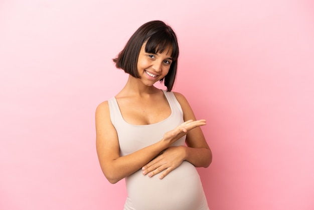 Giovane donna incinta su sfondo rosa isolato che presenta un'idea mentre guarda sorridendo verso