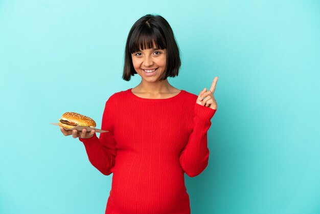 Giovane donna incinta tenendo un hamburger su sfondo isolato che mostra e alzando un dito in segno del meglio