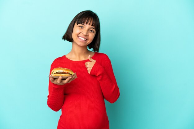 Giovane donna incinta tenendo un hamburger su sfondo isolato dando un pollice in alto gesto