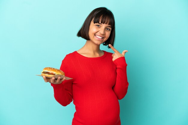 Giovane donna incinta tenendo un hamburger su sfondo isolato dando un pollice in alto gesto