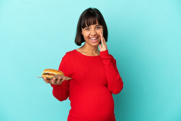 Giovane donna incinta tenendo un hamburger su sfondo isolato gridando con la bocca spalancata