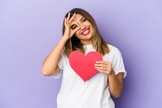 Giovane donna indiana che tiene un cuore di San Valentino isolato eccitato mantenendo il gesto giusto sull'occhio.