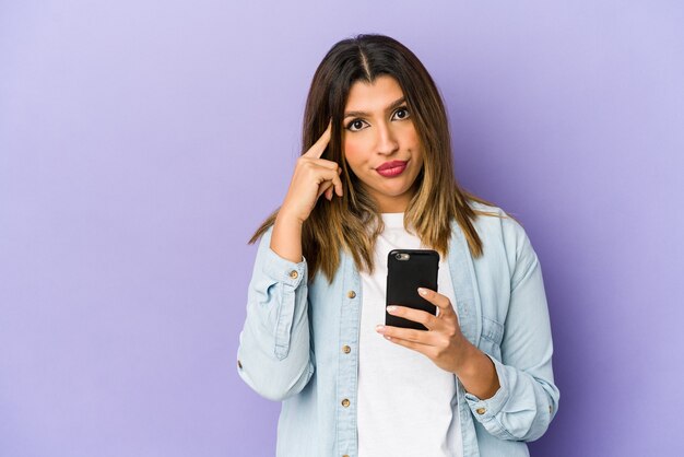 Giovane donna indiana che tiene un tempio di puntamento isolato telefono con il dito, pensando, concentrato su un compito.