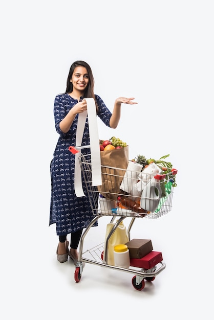 Giovane donna indiana con carrello della spesa o carrello pieno di generi alimentari, frutta e verdura. Isolato Foto a figura intera su muro bianco