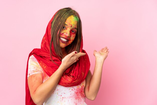 Giovane donna indiana con colorate polveri di holi sul viso sul muro rosa che estende le mani a lato per invitare a venire