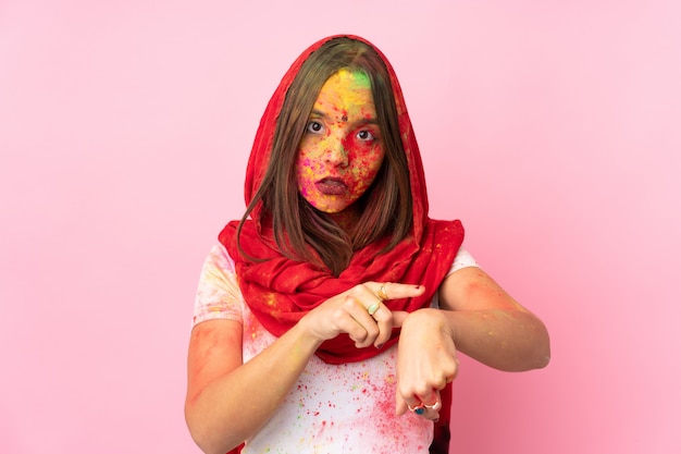 Giovane donna indiana con colorate polveri di holi sul viso sul muro rosa facendo il gesto di essere in ritardo