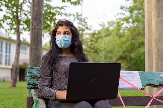 Giovane donna indiana con maschera pensando e utilizzando laptop mentre è seduto con la distanza su una panchina nel parco