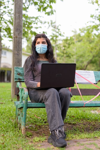 Giovane donna indiana con maschera pensando e utilizzando laptop mentre è seduto con la distanza su una panchina nel parco