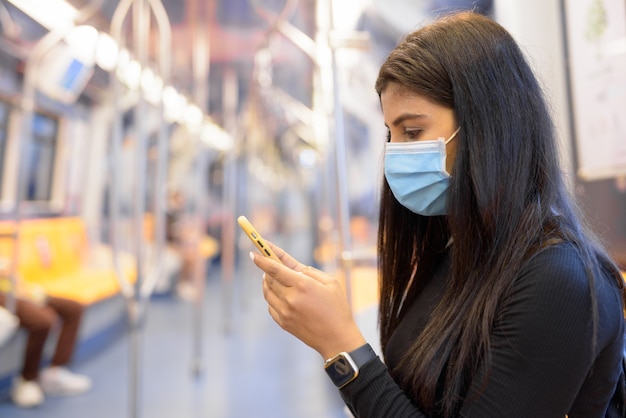 Giovane donna indiana con maschera utilizzando il telefono e seduto con la distanza all'interno del treno