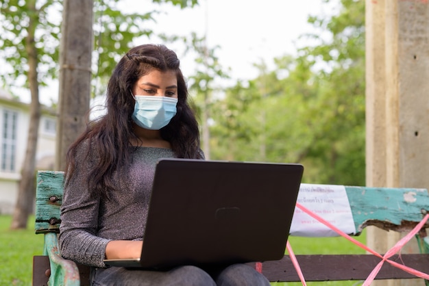 Giovane donna indiana con maschera utilizzando laptop mentre è seduto con la distanza su una panchina nel parco