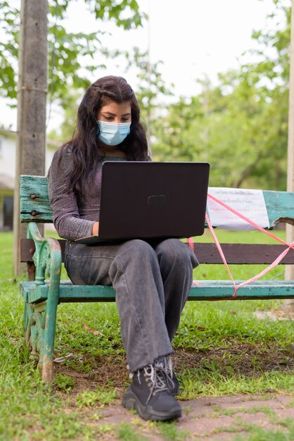 Giovane donna indiana con maschera utilizzando laptop mentre è seduto con la distanza su una panchina nel parco