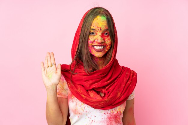 Giovane donna indiana con polveri colorate di holi sul viso sul muro rosa salutando con la mano con espressione felice