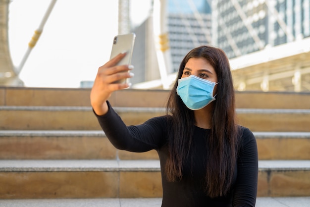 Giovane donna indiana con videochiamata maschera con telefono e seduto dalle scale in città