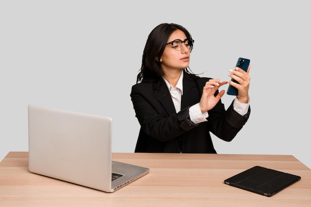 Giovane donna indiana in un tavolo con un computer portatile e un tablet utilizzando un telefono cellulare isolato