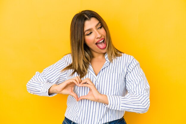 Giovane donna indiana isolata su sfondo giallo sorridente e mostrando una forma di cuore con le mani.