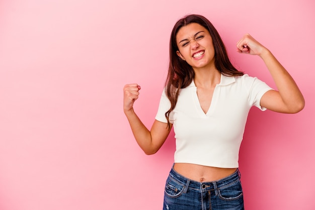 Giovane donna indiana isolata su sfondo rosa alzando il pugno dopo una vittoria, concetto di vincitore.