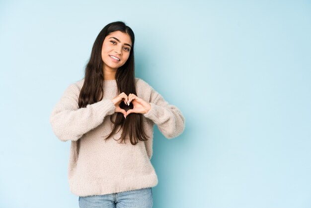 Giovane donna indiana isolata sull'azzurro che sorride e che mostra una forma del cuore con le mani.