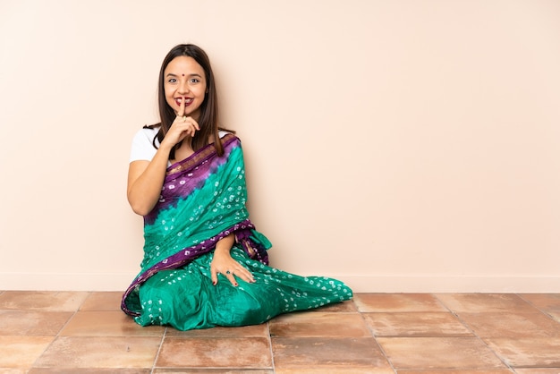 Giovane donna indiana seduta sul pavimento che mostra un segno di silenzio gesto mettendo il dito in bocca