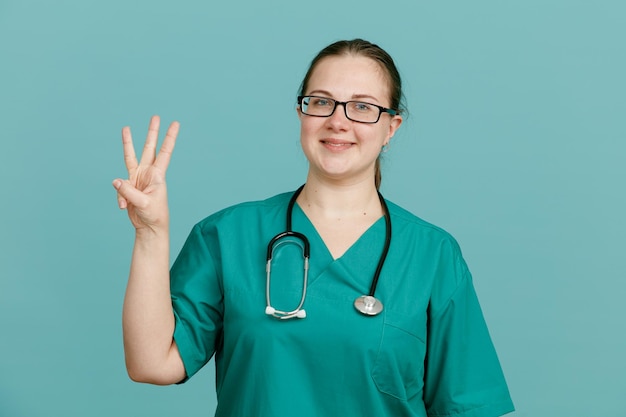 Giovane donna infermiera in uniforme medica con stetoscopio intorno al collo che mostra il numero tre guardando la fotocamera sorridente fiducioso in piedi su sfondo blu