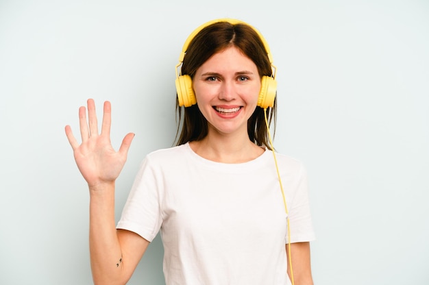 Giovane donna inglese che ascolta la musica isolata su sfondo blu sorridente allegro che mostra il numero cinque con le dita.