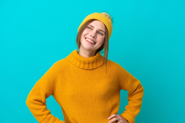 Giovane donna inglese che indossa una giacca invernale isolata su sfondo blu in posa con le braccia all'anca e sorridente