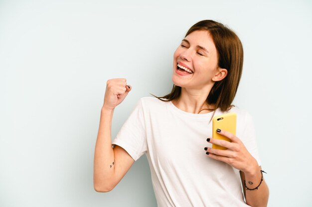 Giovane donna inglese che tiene il telefono cellulare isolato su sfondo blu alzando il pugno dopo un concetto di vincitore della vittoria