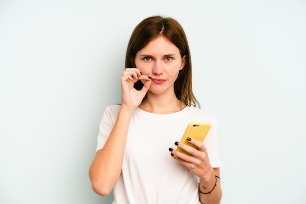 Giovane donna inglese che tiene il telefono cellulare isolato su sfondo blu con le dita sulle labbra mantenendo un segreto.