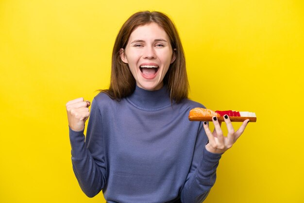 Giovane donna inglese che tiene sashimi isolato su sfondo giallo che celebra una vittoria in posizione di vincitore