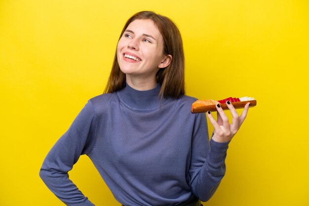 Giovane donna inglese che tiene sashimi isolato su sfondo giallo in posa con le braccia all'anca e sorridente