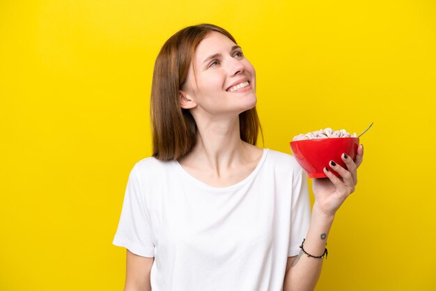 Giovane donna inglese che tiene tazza di caffè isolata su sfondo giallo guardando in alto mentre sorride