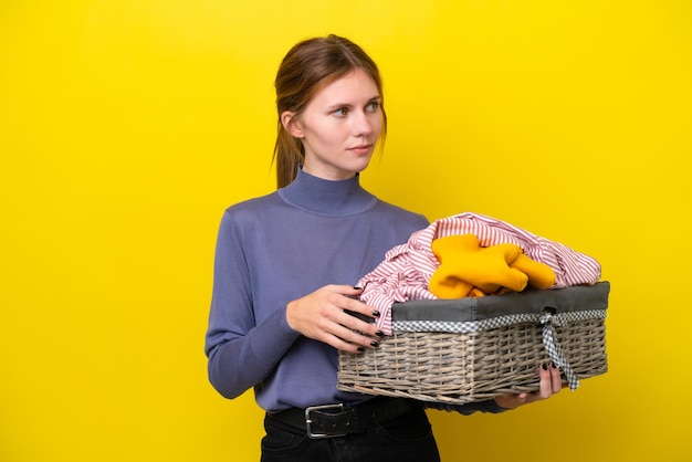 Giovane donna inglese che tiene un cesto di vestiti isolato su sfondo giallo che guarda al lato