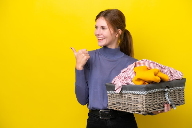 Giovane donna inglese che tiene un cesto di vestiti isolato su sfondo giallo che indica il lato per presentare un prodotto