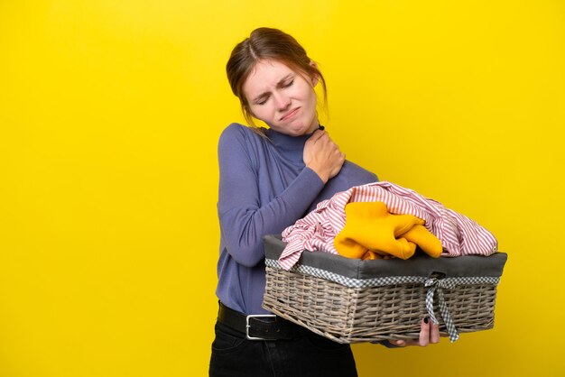 Giovane donna inglese che tiene un cesto di vestiti isolato su sfondo giallo che soffre di dolore alla spalla per aver fatto uno sforzo