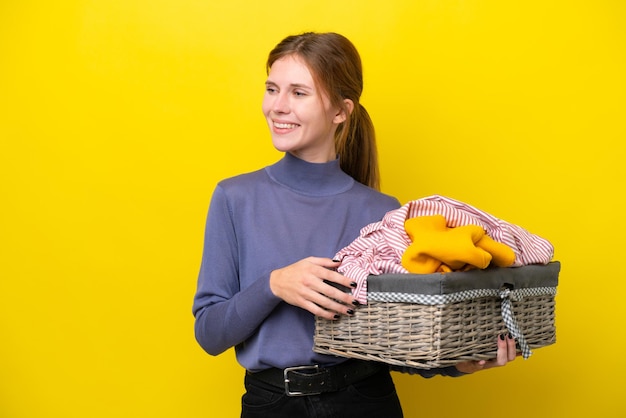 Giovane donna inglese che tiene un cesto di vestiti isolato su sfondo giallo guardando al lato e sorridente