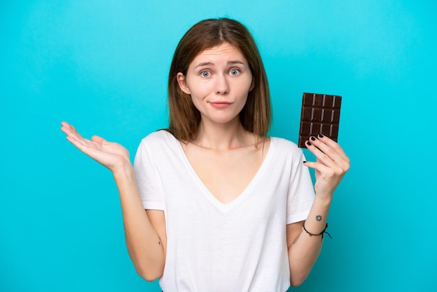 Giovane donna inglese con cioccolato isolato su sfondo blu che ha dubbi mentre alza le mani