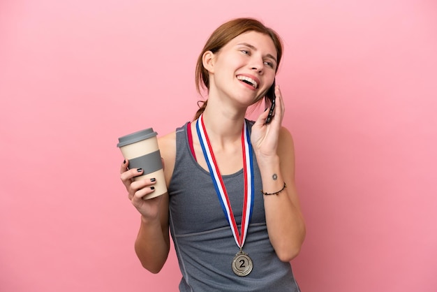 Giovane donna inglese con medaglie isolate su sfondo rosa che tiene caffè da portare via e un cellulare