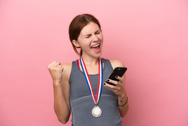 Giovane donna inglese con medaglie isolate su sfondo rosa con telefono in posizione di vittoria