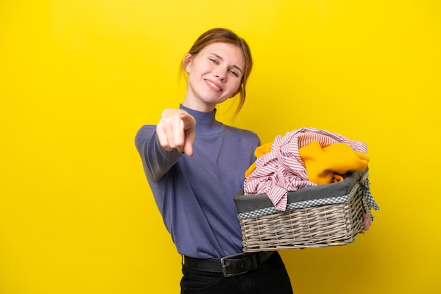 Giovane donna inglese in possesso di un cesto di vestiti isolato su sfondo giallo che punta davanti con felice espressione