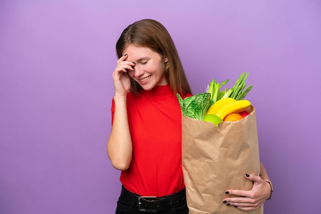 Giovane donna inglese in possesso di un sacchetto della spesa isolato su sfondo viola ridendo