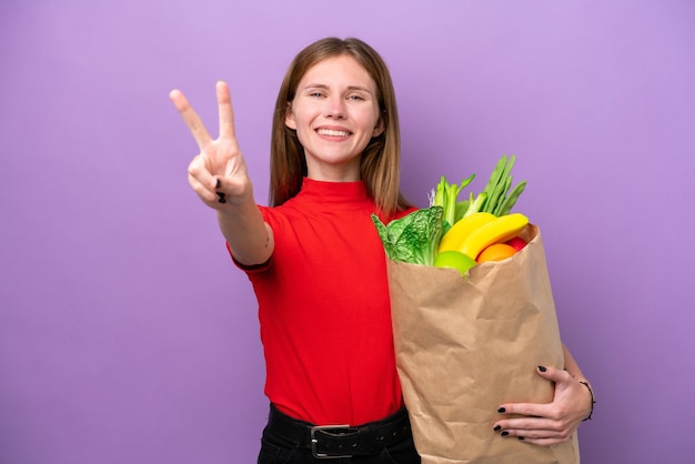 Giovane donna inglese in possesso di un sacchetto della spesa isolato su sfondo viola sorridente e mostrando il segno di vittoria