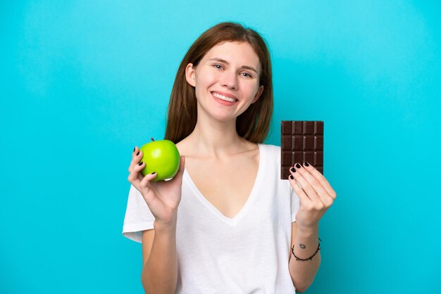 Giovane donna inglese isolata su sfondo blu che prende una tavoletta di cioccolato in una mano e una mela nell'altra