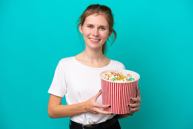 Giovane donna inglese isolata su sfondo blu con in mano un grande secchio di popcorn