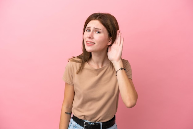 Giovane donna inglese isolata su sfondo rosa che ascolta qualcosa mettendo la mano sull'orecchio