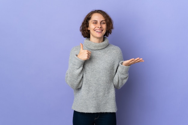 Giovane donna inglese isolata su sfondo viola che tiene copyspace immaginario sul palmo per inserire un annuncio e con i pollici in su