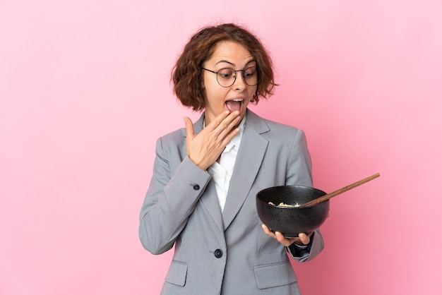 Giovane donna inglese isolata sulla parete rosa con sorpresa e espressione facciale scioccata mentre si tiene una ciotola di spaghetti con le bacchette