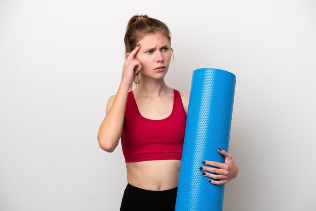 Giovane donna inglese sportiva che va a lezioni di yoga mentre tiene un tappetino isolato su sfondo bianco con dubbi e pensare